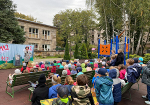 zdjęcie wykonane podczas teatrzyku w naszym ogrodzie przedszkolnym, dzieci siedzą na dużych drewnianych ławkach przodem do przedszkola i przodem do aktorów, w oddali widać drzewa