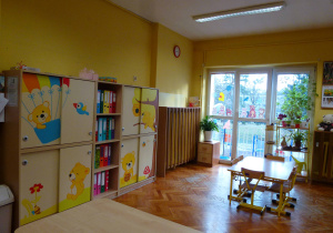 część sali 4-latków widocznej od wejścia, żółte ściany, regał na pomoce dydaktyczne, na zasuwanych szafkach znajdują się ozdoby-żółte misie, na dalszym planie widać okna balkonowe oraz jeden stolik i krzesła