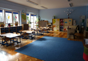 sala 5-latków, z niebieskimi ścianami i niebieskim dywanem, widok na okna, pod którymi stoją stoliki z krzesłami oraz szuflady na osobiste rzeczy dzieci, pod oknem balkonowym w rogu sali znajduje się kącik przyrodniczy z kwiatami na stojakach