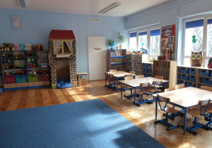 sala 5-latków, widok na jej tylną część, po prawej stronie okna, na lewo od okien wejście do schowka, dalej na lewo domek papierowy stojący pod ścianą, a najbardziej po lewej stronie stoją regały z zabawkami dla dzieci, na bliższym planie stoją stoliki z krzesłami