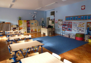 sala 5-latków, z niebieskimi ścianami i niebieskim dywanem, widok na przednią część sali, widać stoliki, krzesła, tablicę dydaktyczną, biurko nauczycielki i kolorowe regały z chmurkami, jeżykiem i drzewem.