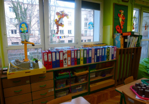sala 6-latków z zielonymi ścianami i zielonymi roletami, na regale pod oknem ustawione są segregatory dzieci, w których trzymają swoje prace, obok na pólkach stoją zabawki, a niżej ustawione są na półkach pudełka z klockami