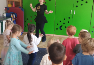 Na zdjęciu widać grupę dzieci stojących tyłem lub bokiem i w oddali na tle zielonej szafy nauczycielkę tańca, która pokazuje dzieciom ruchy taneczne.