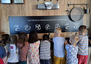 Na zdjęciu widać dzieci, które myją ręce. Powyżej na ścianie narysowana i opisana instrukcja mycia rąk.