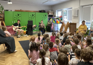 Sala rytmiczna w szerszym planie, pani śpiewa, pan gra na gitarze, widać zainteresowanie dzieci koncertem.