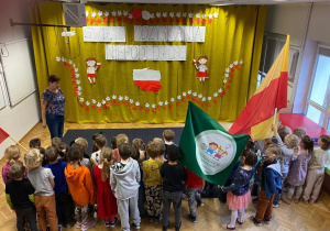 Na zdjęciu widać stojace dzieci oraz panią. Jest kotara z symbolami narodowymi i napisem "Święto odzyskania niepodległości". Widać też flagę Warszawy i chorągiew przedszkola nr 61.