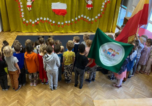 Dzieci stoją i patrzą na kotarę z symbolami narodowymi.