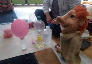 Widać różowy balon, butelki z różnymi substancjami i maskotkę Labolka.