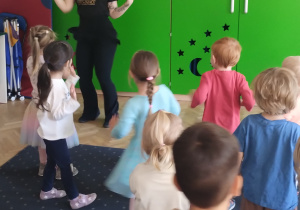 Na zdjęciu widać grupę dzieci stojących tyłem lub bokiem i w oddali na tle zielonej szafy nauczycielkę tańca, która pokazuje dzieciom ruchy taneczne.