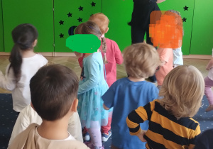 Na zdjęciu widać grupę dzieci stojących tyłem lub bokiem i w oddali na tle zielonej szafy nauczycielkę tańca, która pokazuje dzieciom ruchy taneczne.