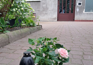 Na pierwszym planie znicz i kwiaty, w perspektywie widoczne drzwi, obok których znajduje się tablica pamiątkowa.