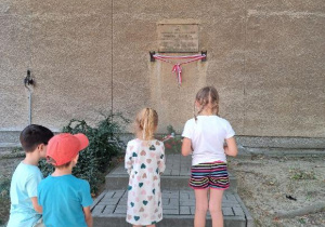 Dzieci stoja przed tablicą upamiętniajacą Powstańców Warszawskich z Ochoty.