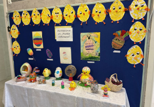 Na granatowej tkaninie na ścianie wiszą obrazki namalowane lub wyklejane przez dzieci przedstawiające piaanki.Poniżej na stolikach przykrytych białym obrusem stoją wykonane przez dzieci jajka pisanki.